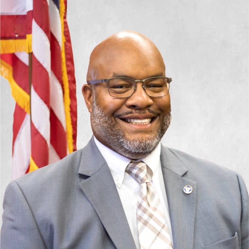 Headshot of a black man with a shaved head, glasses and short cropped beard wearing a grey suit and plaid Tie with a US flag in the background